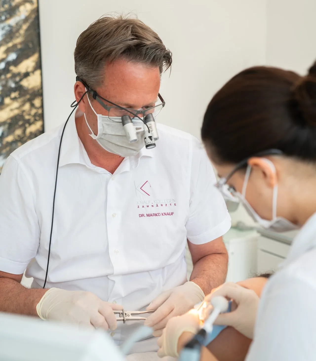 Dr. Marko Knauf bei einer Behandlung mit Lupenbrille, an seiner Seite eine Assistentin